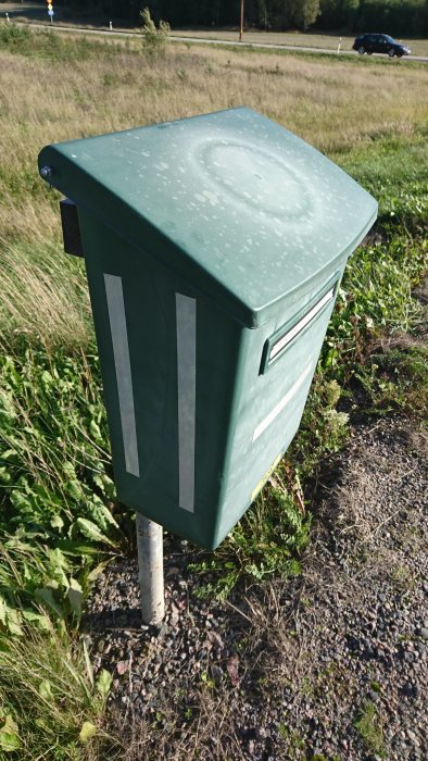 Grön brevlåda med stängt lock vid en vägkant, används för att diskutera fjädersäkring mot vind.