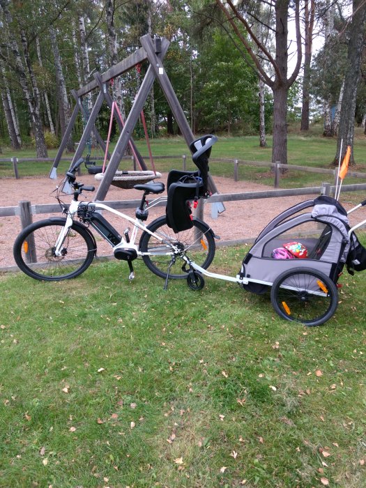 Elcykel med barnstol och kopplad cykelvagn står i en parkmiljö.