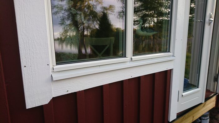 Nyinstallerade vita fönsterbleck och bleck runt en pardörr på en röd trävägg av ett hus med spegling av träd i fönstret.