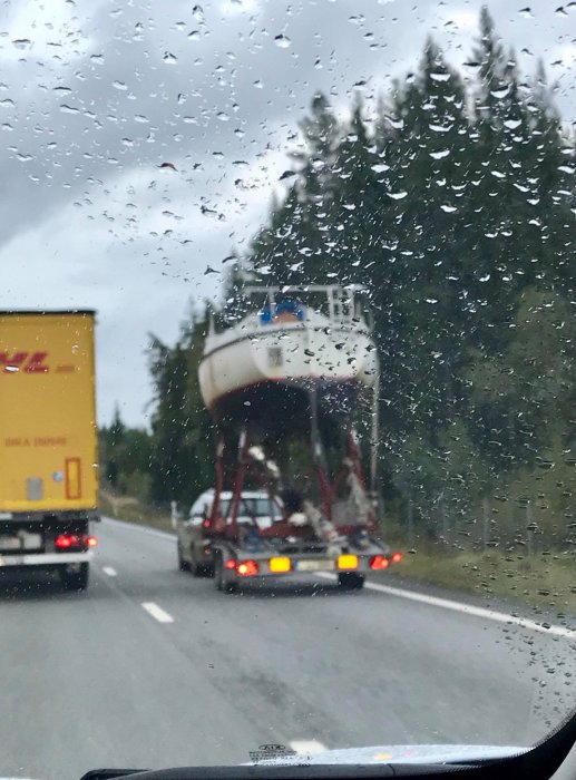Båt transporterad på släpvagn bakom en lastbil på regnig väg, sett genom ett blött bilfönster.