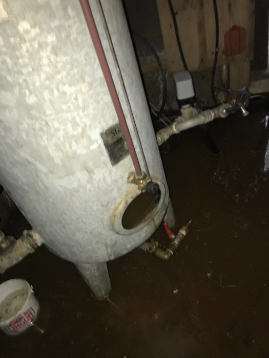 Hydrofor med öppen front, vattenläckage, rör och verktyg på ett smutsigt golv.
