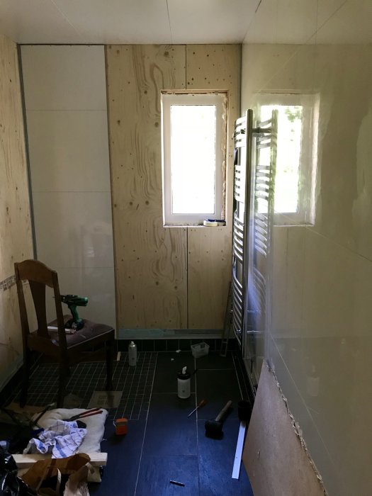 Ofärdigt badrum med plywood och delvis monterade väggskivor, verktyg och byggmaterial på golvet.