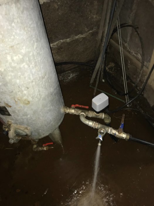 Hydrofor med pågående vattenpåfyllning och öppen kran som visar vattentrycket, rör och kablar i bakgrunden.