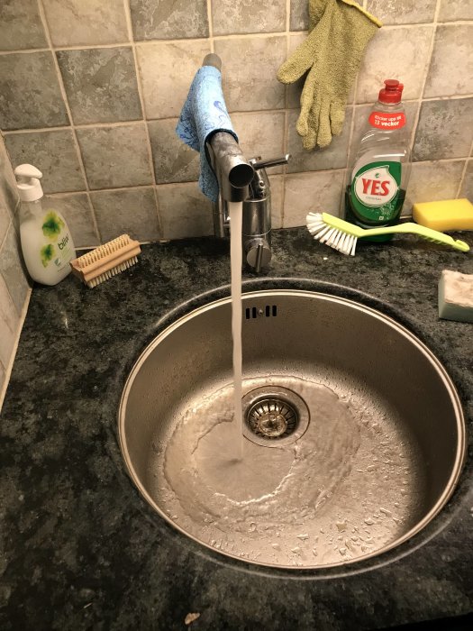 Köksdiskho med rinnande vatten, rengöringsmedel och diskborste på bänken.