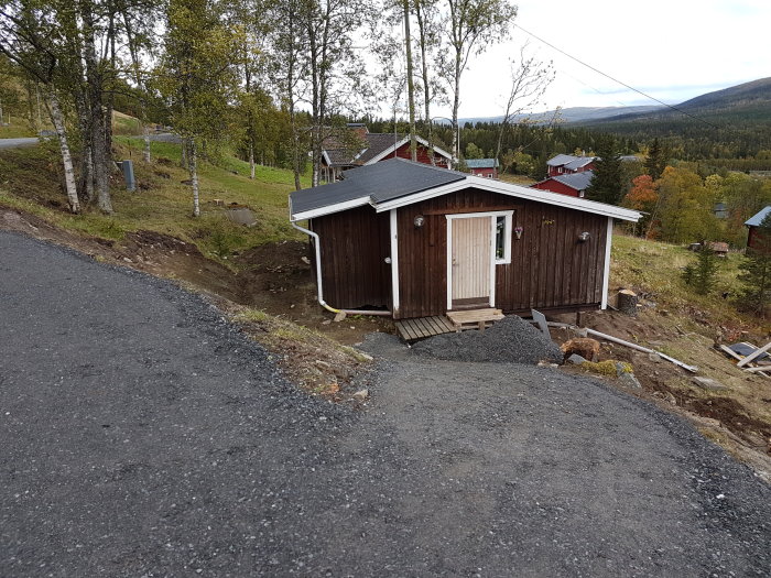 Nybyggd grusväg som leder upp till ett brunt trähus med vita knutar, omgiven av höstfärgad natur.