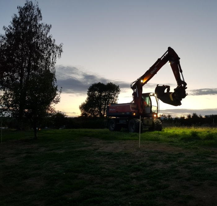 Grävmaskin i arbete vid skymningen på en gräsmark, med silhuetter av träd och en färgrik himmel i bakgrunden.