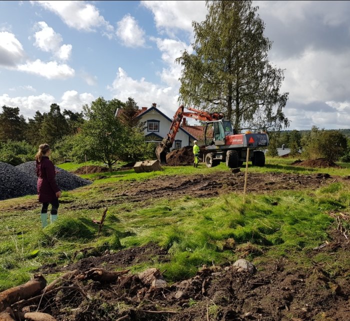 Grävarbete vid en trädgård med grävmaskin och kvinna som letar stenar, hus och träd i bakgrunden.