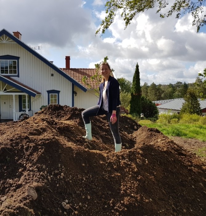 Glad kvinna står på en hög med grävd jord framför ett hus med glada miner under grävarbeten.