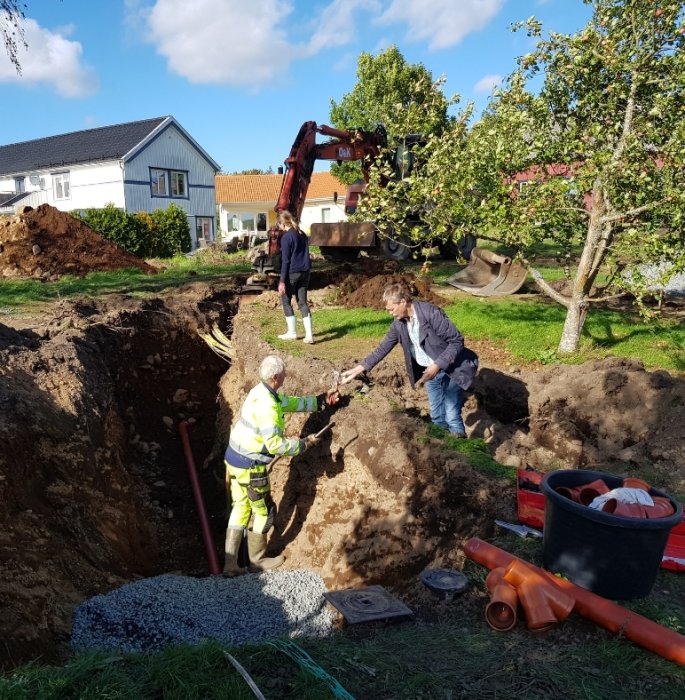 Grävarbete i trädgård med grävmaskin och flera personer som deltar, inklusive en äldre man och kvinna som plockar stenar.