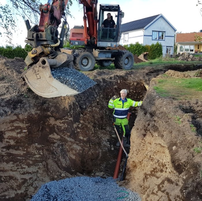 Äldre man i arbetskläder står i en grävd grop bredvid en grävmaskin och grushög framför ett hus.