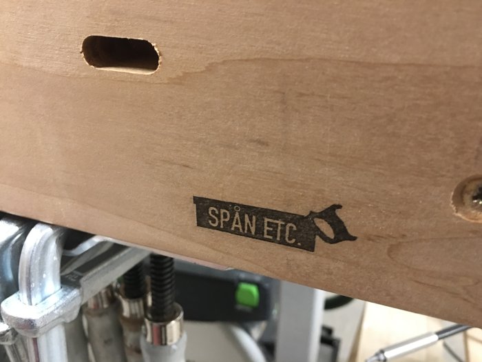 Närbild på arbetsbord med utskurna hål och svart text "SPÅN ETC." bredvid ett symboliskt sågverktyg.