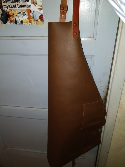 Läderförkläde med remmar hängandes på en vit dörr, designat för snickeriarbete.