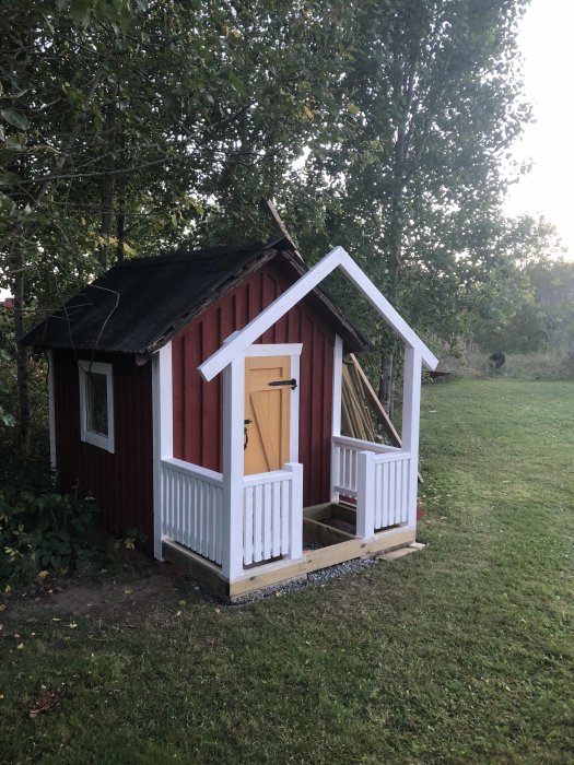 Ny målad farstukvist på röd lekstuga med vit staketsektion omgiven av grönt gräs och träd.