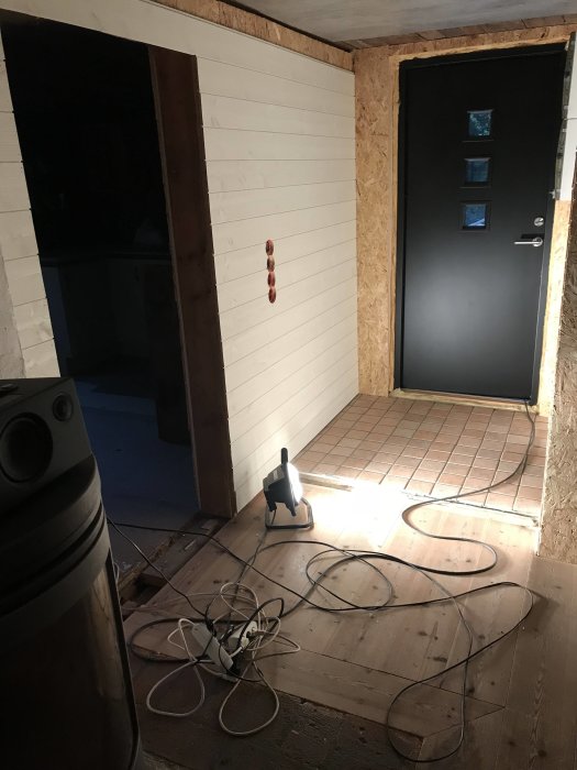 Renoverad hall med ny svart ytterdörr, vita väggar och oskyddad väggpanel väntar på golvläggning, med arbetslampa och kablar på golvet.