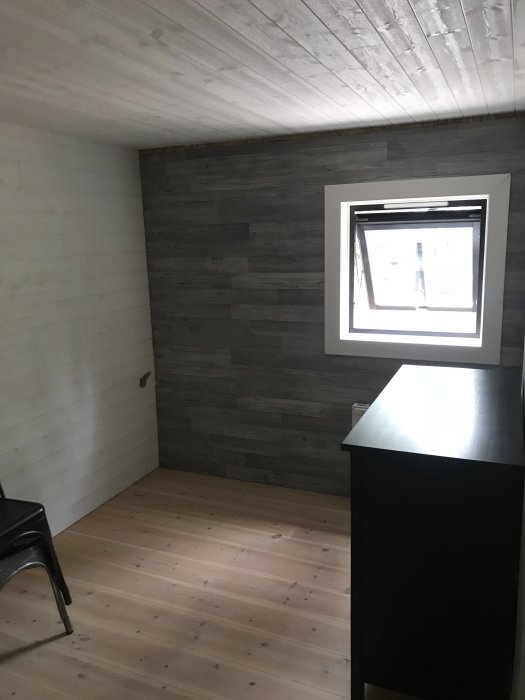 Renoverad hall med nyinstallerad grå väggpanel, vitmålade väggar och tak, trägolv och ett svart skåp.