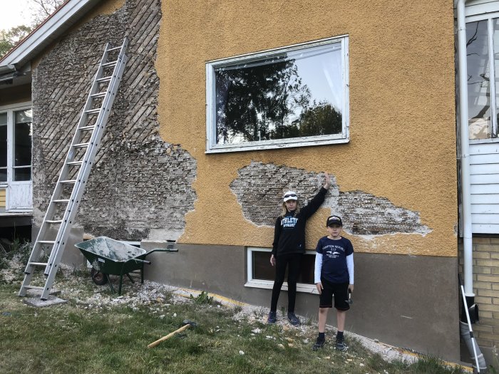 Två barn står framför ett hus med delvis borttagen ytterputs och byggmaterial synliga, stege och skottkärra i förgrunden.