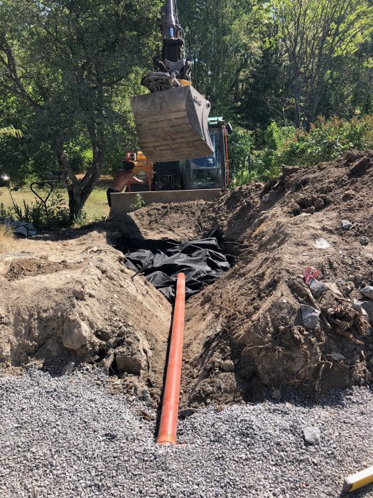 Grävmaskin ovanför nygrävd schaktbotten med grus, svart fiberduk och orange dräneringsrör som leder mot stenkista.