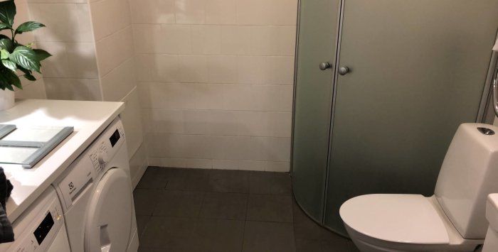 En bild av ett badrum med vita kaklade väggar, mörkt klinkergolv, tvättmaskin, toalett och grön duschvägg.