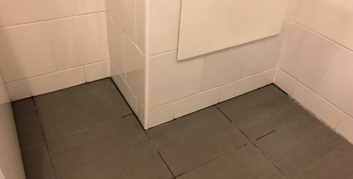 Hörn av badrum med kaklade väggar och klinkergolv som visar fuktmarkering i fogar.