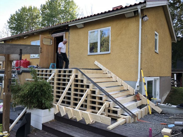 Nykonstruerad trätrappa under uppbyggnad utanför ett gult hus med en person som arbetar.