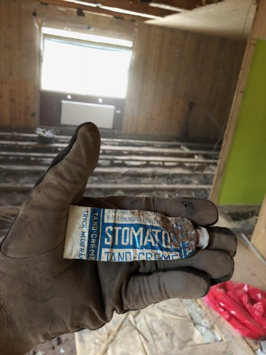 Hand med skyddshandske håller gammal tandkrämstub från 1951 i renoveringsrum med nakna bjälkar.