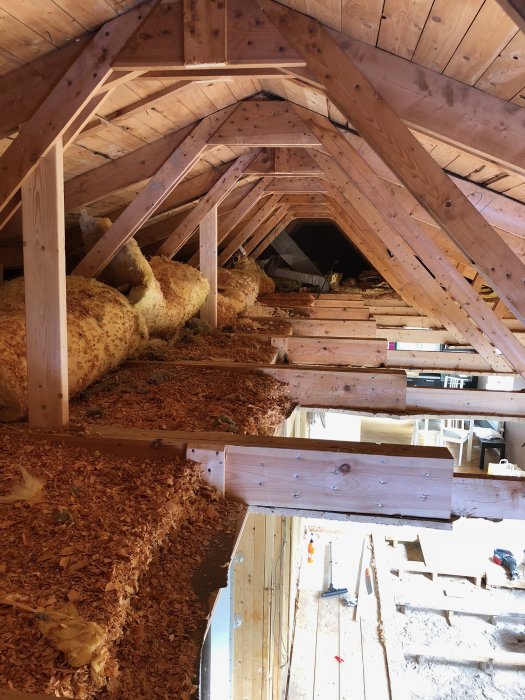 Takstolar i trä och en stor limträbalk som stöd, med uppriven isolering och spån på golvet i en husattik under renovering.