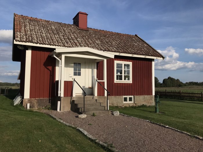 Rödmålat torp med vitkantade fönster och dörr, grusgång och enkel grön handpump framför huset.