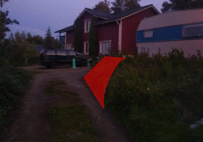 Ogräsigt område med en husvagn och en skrotbil mellan två hus i skymningen, markerat för borttagning.