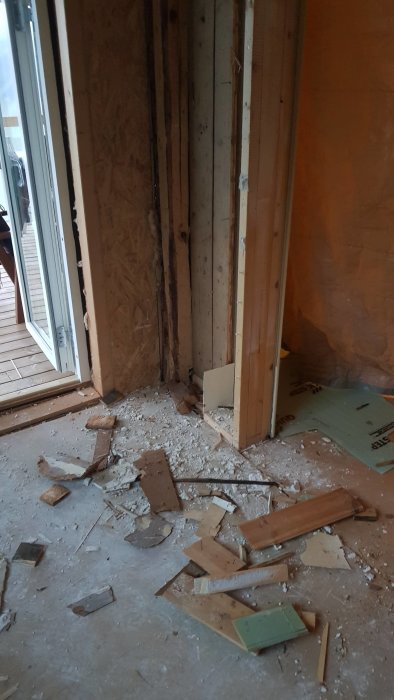 Hörn av ett rum under renovering med en stående trästock bultad vid yttervägg och spillror på golvet.