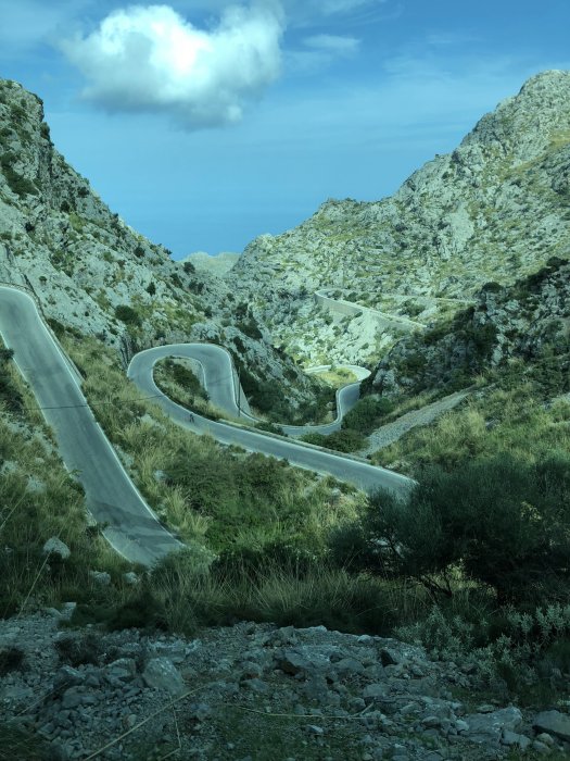 Vindlande bergsväg i Mallorca med kuperat landskap och gröna buskar under en blå himmel med moln.
