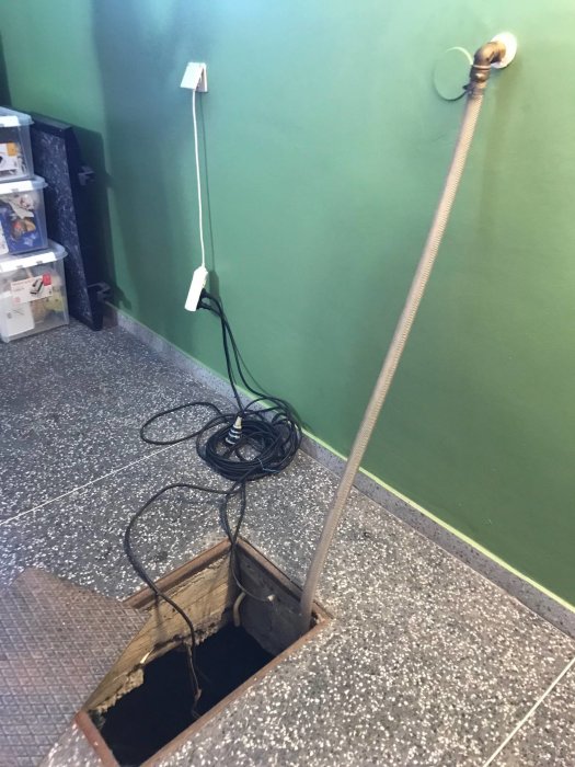 Öppen brunnslock i ett garage med synlig pump och slang, oordnad kabelhög och strömkabel på väggen.