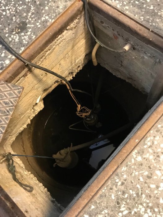 Rund betongbrunn med pump och slangar i ett garage, synliga genom öppen lucka i golvet.