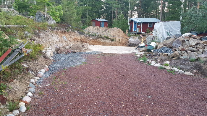 Förberedd byggplats för garage med jämnad berghäll och grus, omgiven av natur och röda stugor.