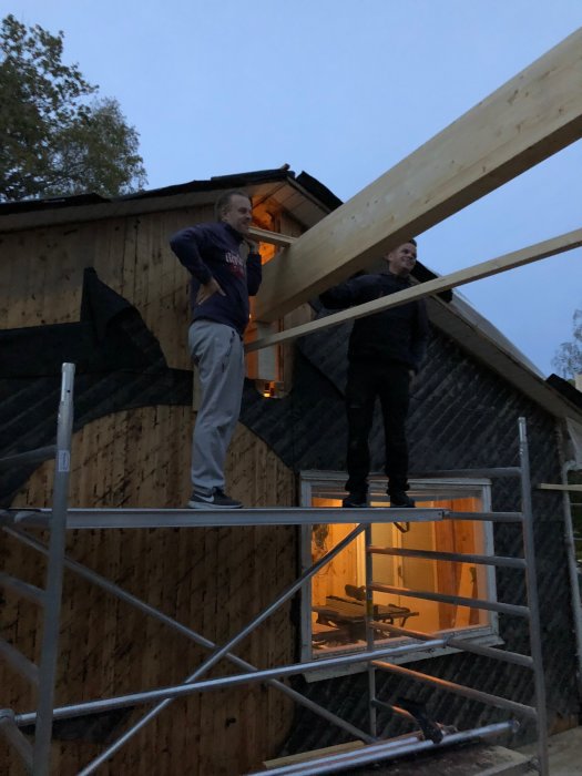 Två personer lyfter en stor limträbalk på en byggarbetsplats under kvällstid.