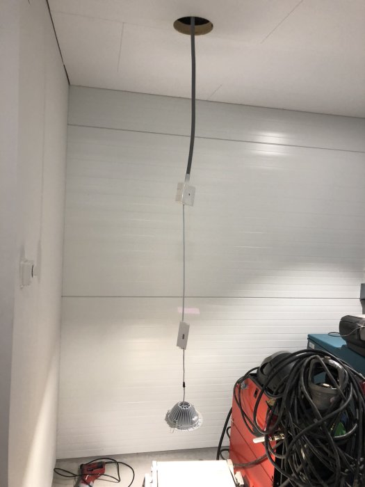 En lampa i taket med hängande kabel och icke-monterad lampskärm mot en vit vägg.