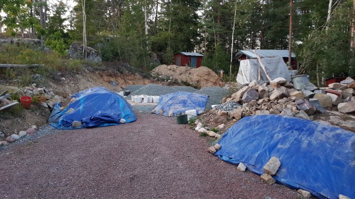 Byggplats med grus- och sandhögar under presenningar, omgiven av berg och skog.