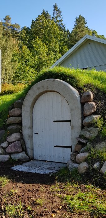 Vitmålad rundad dörr i en gräsbeklädd kulle med stenar runt omkring och en träbyggnad i bakgrunden.