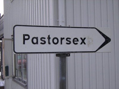 Vägskylt med texten "Pastorsex" pekande åt höger framför en vågrät ribbad vit vägg.