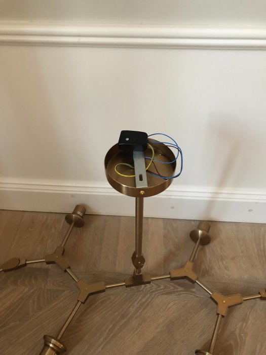 Omonterad taklampa i guldton med synliga kablar ligger mot vit vägg och parkettgolv, i väntan på upphängning.