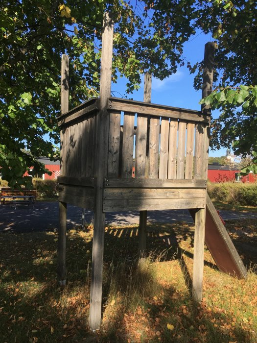 Ett slitet trälektorn med rutschkana på en gräsbevuxen yta, omringat av träd och löv på marken.