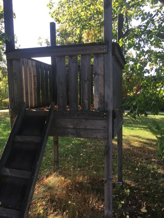 Ett slitet trälekstorn med rutschkana på en övergiven lekplats omgiven av gräs och träd i dagsljus.