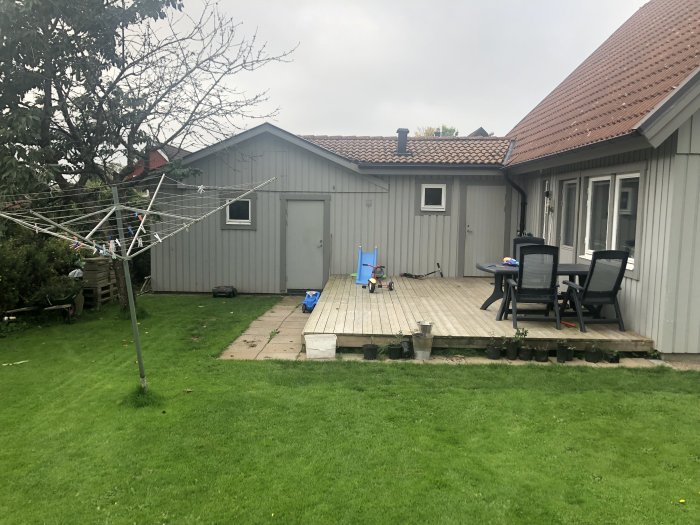 Trädgård med trädäck intill husets baksida, leksaker på gräs, torkställning och höjdskillnad till garage.