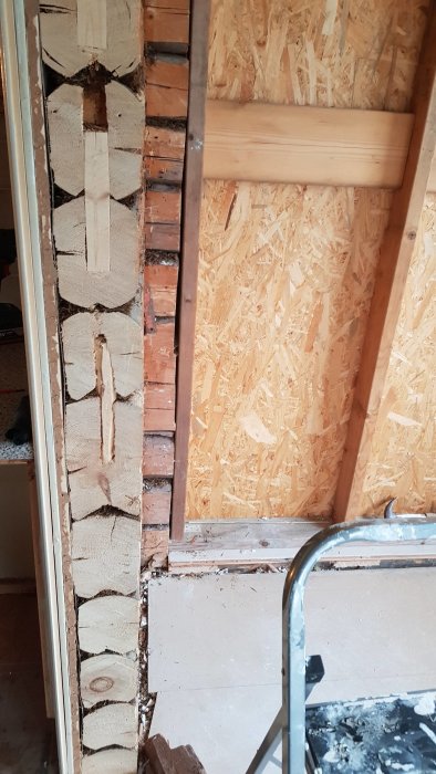 Väggsektion med synliga timmerstockändar intill OSB-skiva och isolering i en byggmiljö, fotograferat inifrån.