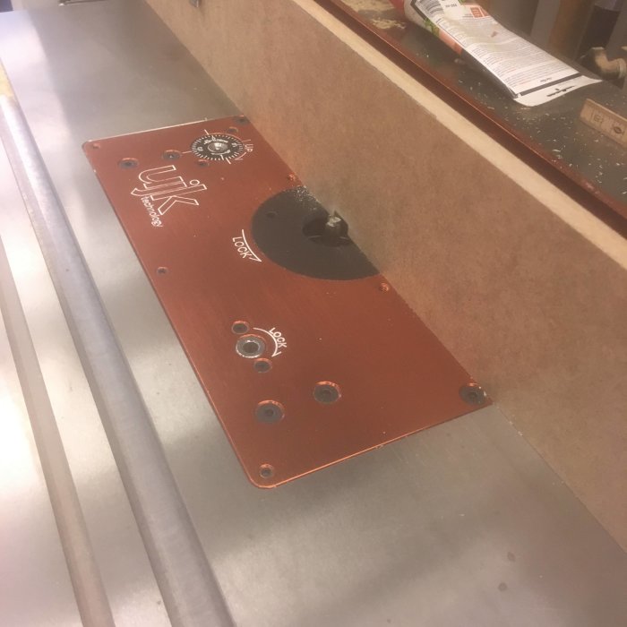 Fräsverktyg med anhåll och fasfräs installerat på ett arbetsbord för träbearbetning, synliga spån och en tidning i bakgrunden.