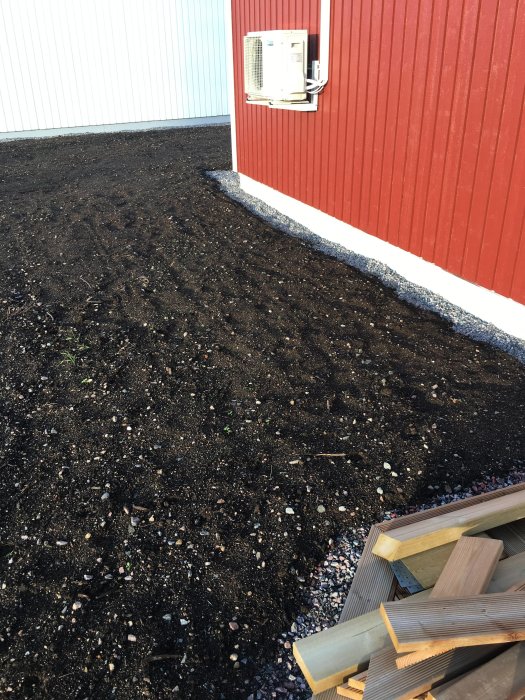 Matjord längs en röd husvägg med synliga småstenar och en bit byggnadsmaterial i förgrunden.
