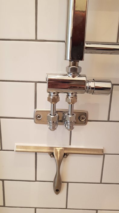 Kromad duschblandare monterad på vit kakelvägg ovanför metallisk rakbladsskrapa.