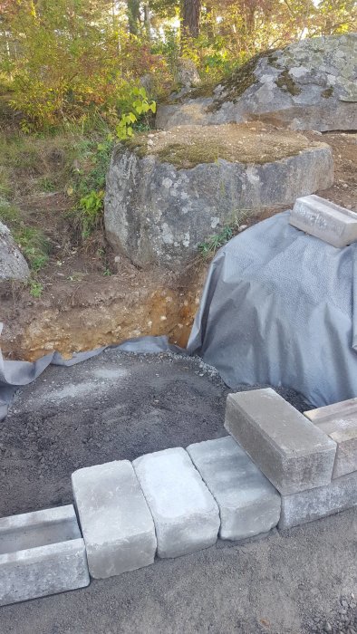 Byggprocess av trappsteg nära en stor sten, med geotextil och delar av stödmur synlig.