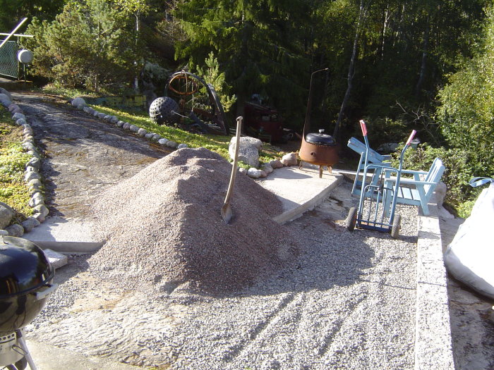 Sten och sand upplagda på en gårdsplan i skogsmiljö, verktyg och byggmaterial synliga.