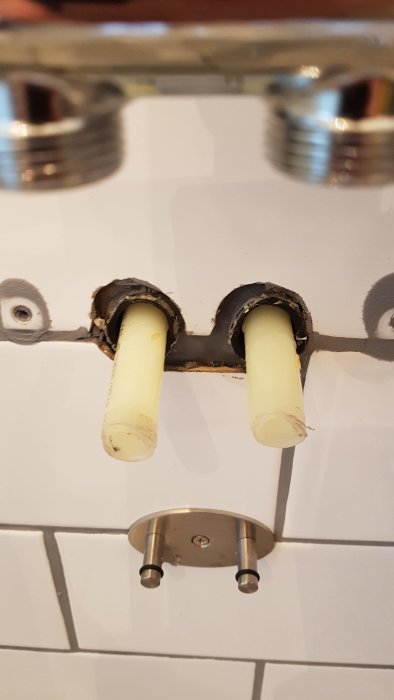 Två plaströr sticker ut från en kakelvägg med en demonterad metallfäste för badrumsutrustning.