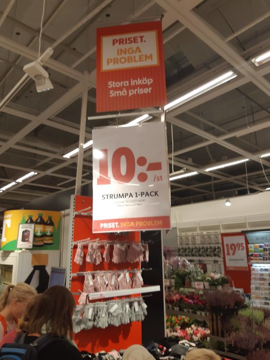 Prisskylt i butik som annonserar "STRUMPA 1-PACK" för 10 kronor med bakgrund av affärshyllor och kunder.